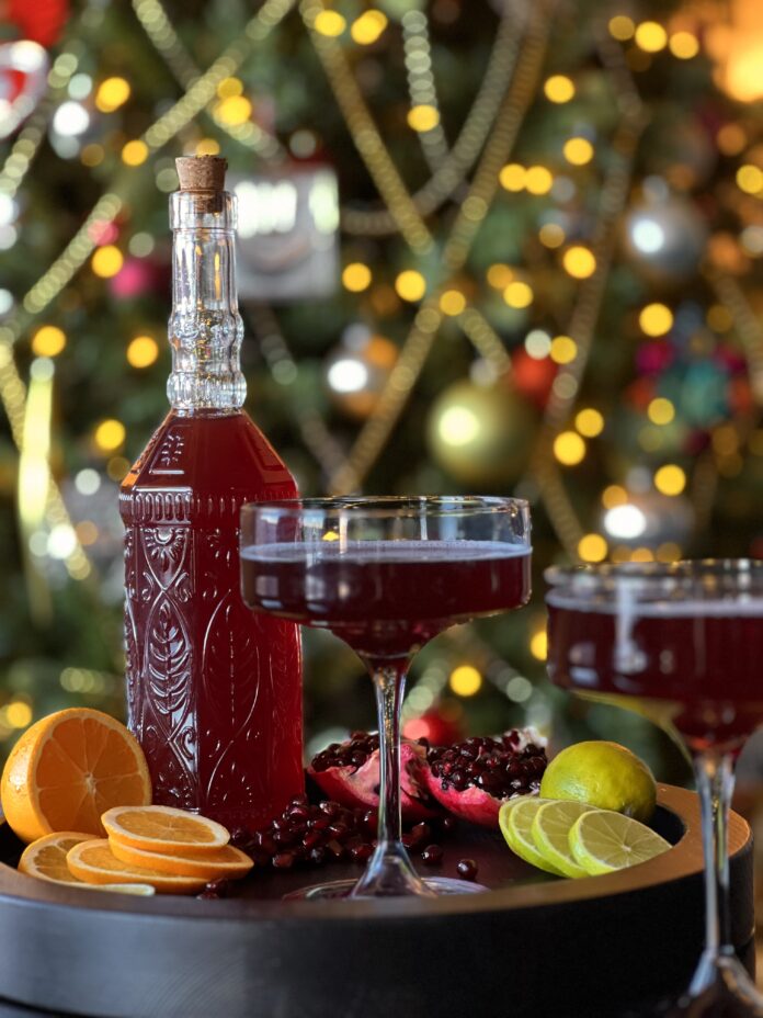 Good Luck Sparking Pomegranate Cosmo A fancy bottle with a deep red Good Luck Sparkling Pomegranate Cosmo on a tray with 2 cocktail glasses, orange slices, an open pomegranate, and lemon slices in front of a blurred Christmas tree.