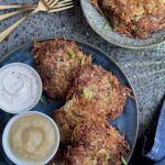 A round blue platter with 5 potato latkes , two small bowls with sour cream and applesauce, 3 gold forks, and a smaller lighter blue plate with 3 smaller latkes, and a blue linen.