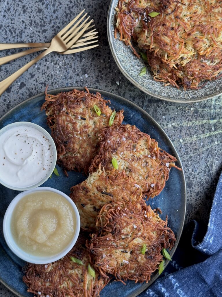 A round blue platter with 5 potato latkes , two small bowls with sour cream and applesauce, 3 gold forks, and a smaller lighter blue plate with 3 smaller latkes, and a blue linen.