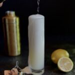 The Gin Fizz with Rizz: A tall, thin highball glass with a cocktail topped with champagne on a black background with a gold cocktail shaker, lemon, and the bottle cork as styling elements.
