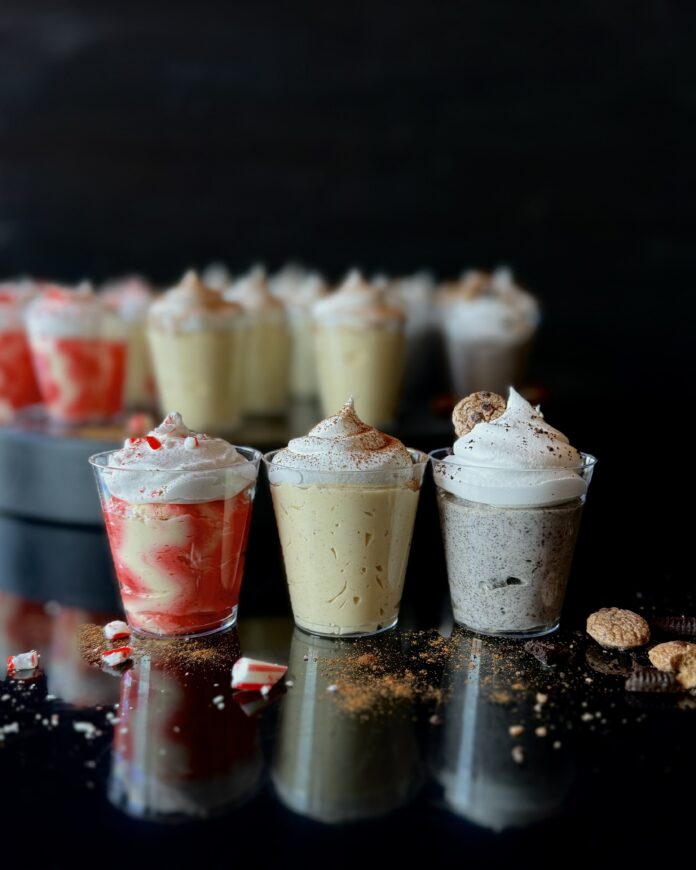 A trio of Christmas and NYE festive pudding shots topped with whipped cream, lined up in a row on a black reflective surface with a tray filled with pudding shots in the background.
