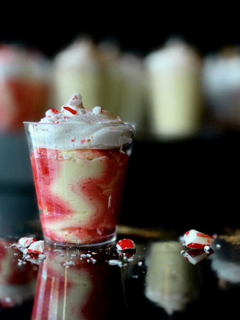 A Candy Cane Forest Pudding Shot with a swirled red and ivory pudding in a small clear cup topped with whipped cream and peppermint pieces sitting on a reflective black surface with blurred pudding shots in the background.