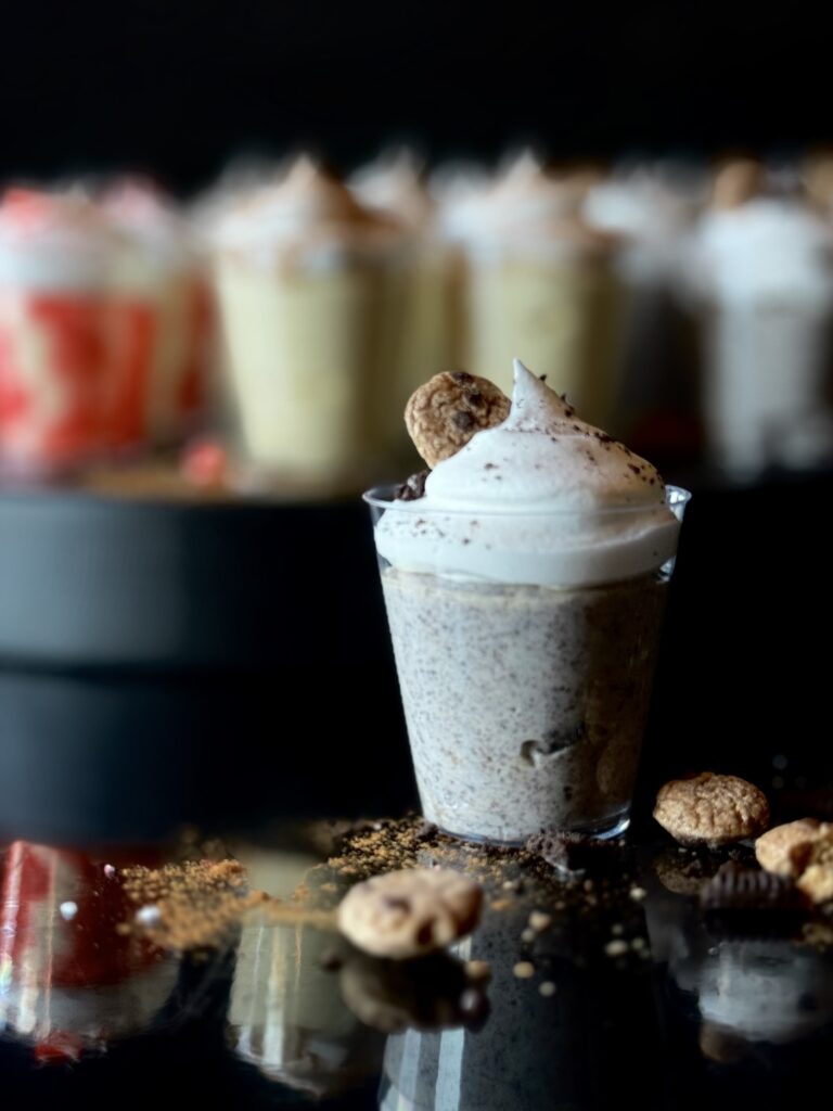 A clear cup with cookie pudding shots topped with whipped cream and garnished with a tiny chocolate chip cookie, sitting on a black reflective surface with a tray of blurred pudding shots in the background.