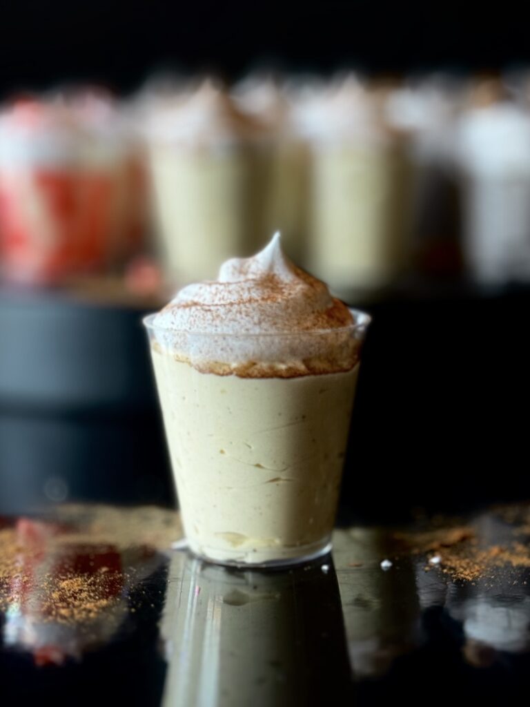 An eggnog pudding shot in a clear cup sitting on a reflective surface with a tray of blurred out pudding shots in the background.