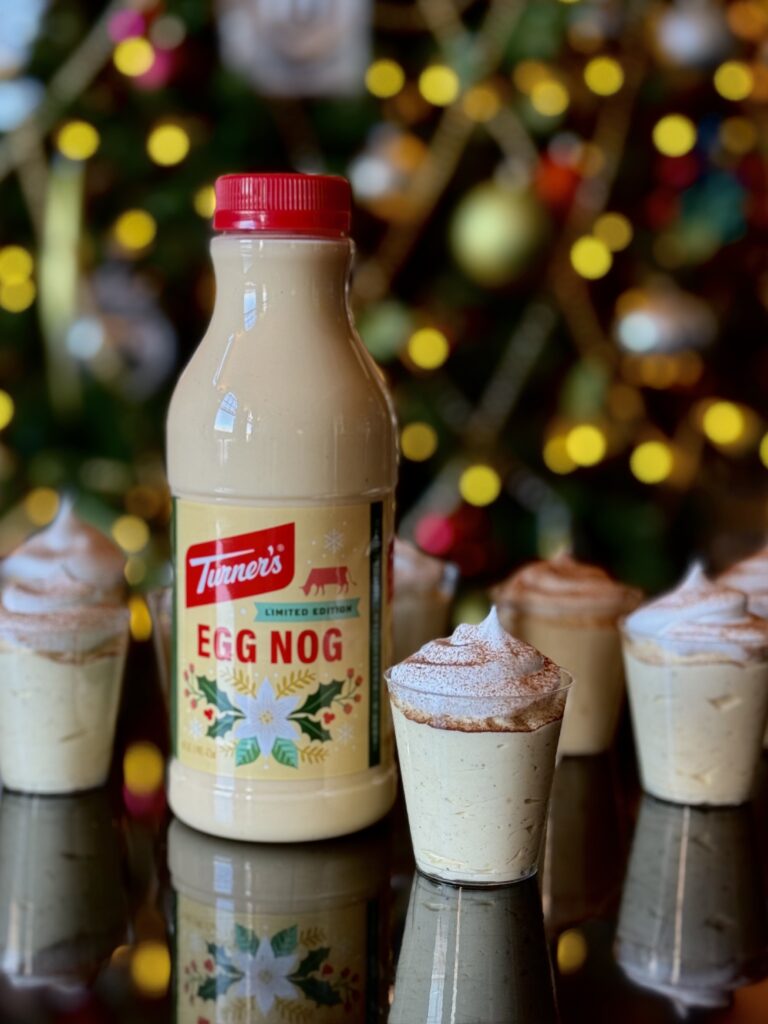 Eggnog Pudding Shots in clear plastic shot glasses with a bottle of Turner's eggnog sitting on a black reflective surface in front of a lit Christmas tree.