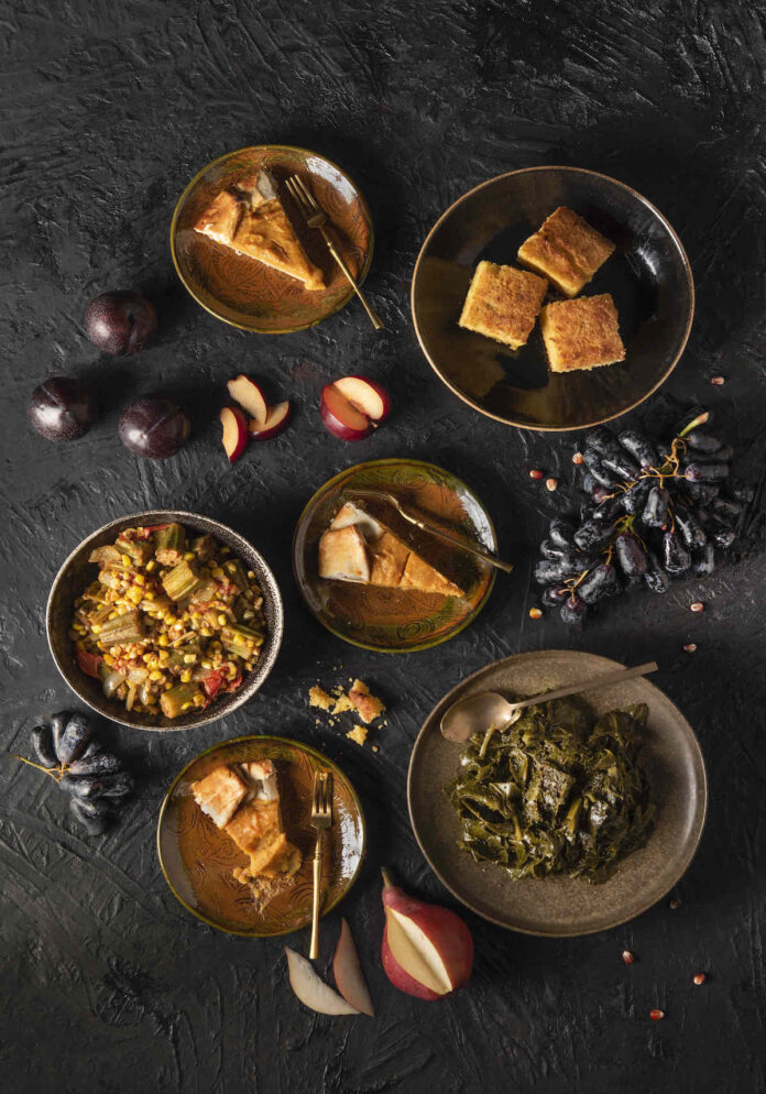 A variety of plates filled with different sides of cornbread, collard greens, and so much more is laid out on a big black table.
