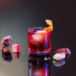 A glass of a red Boulevardier cocktail for Aries topped with an orange twist and big ice cube.