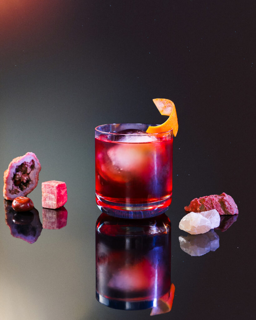 A glass of a red Boulevardier cocktail for Aries topped with an orange twist and big ice cube.