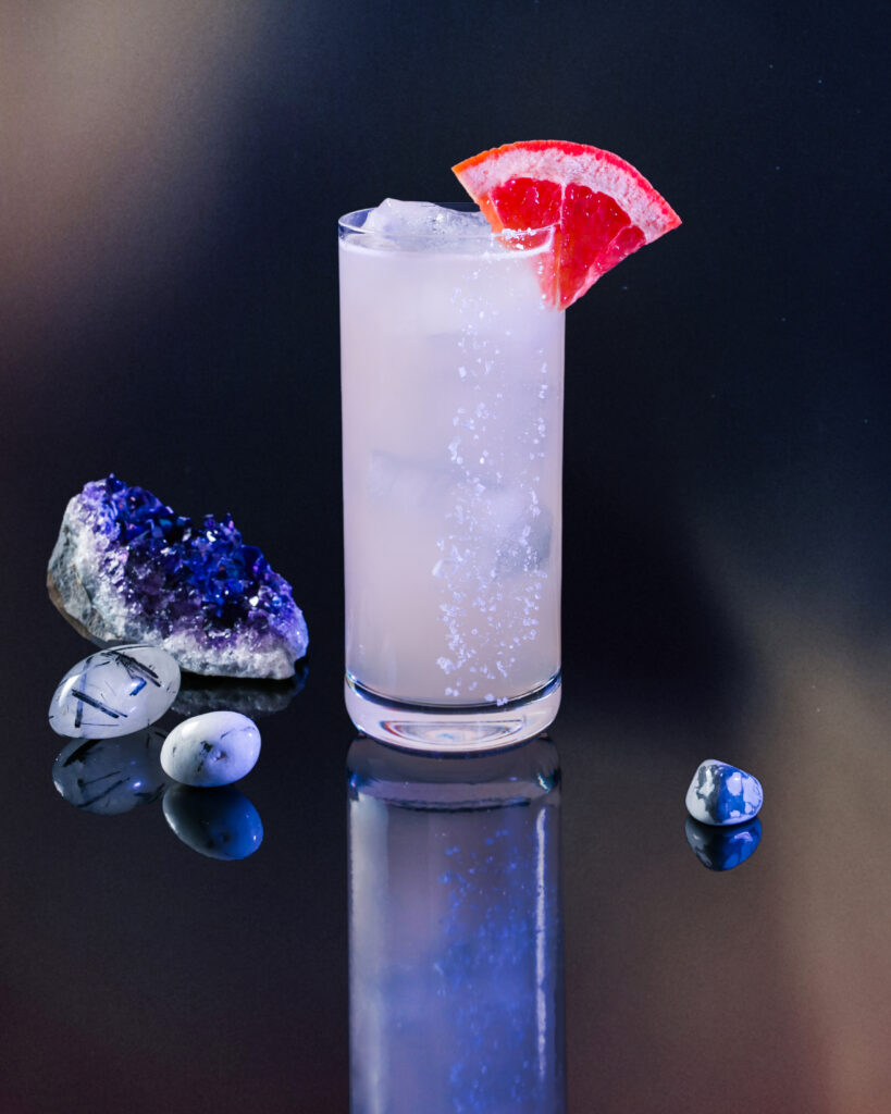 An Aquarius inspired Paloma cocktail filled with white looking liquid and topped with a slice of blood orange.