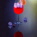 A cocktail for Leo with red liquid in a wine glass topped with an orange slice and surrounded by crystals on the table.
