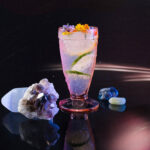 A gin and tonic cocktail for Taurus sits on a black table in a tall glass topped with different citruses and surrounded by geodes.