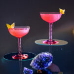 A cocktail for Libra includes tow glasses filled with a pink cosmopolitan with lemon garnish against a black background.
