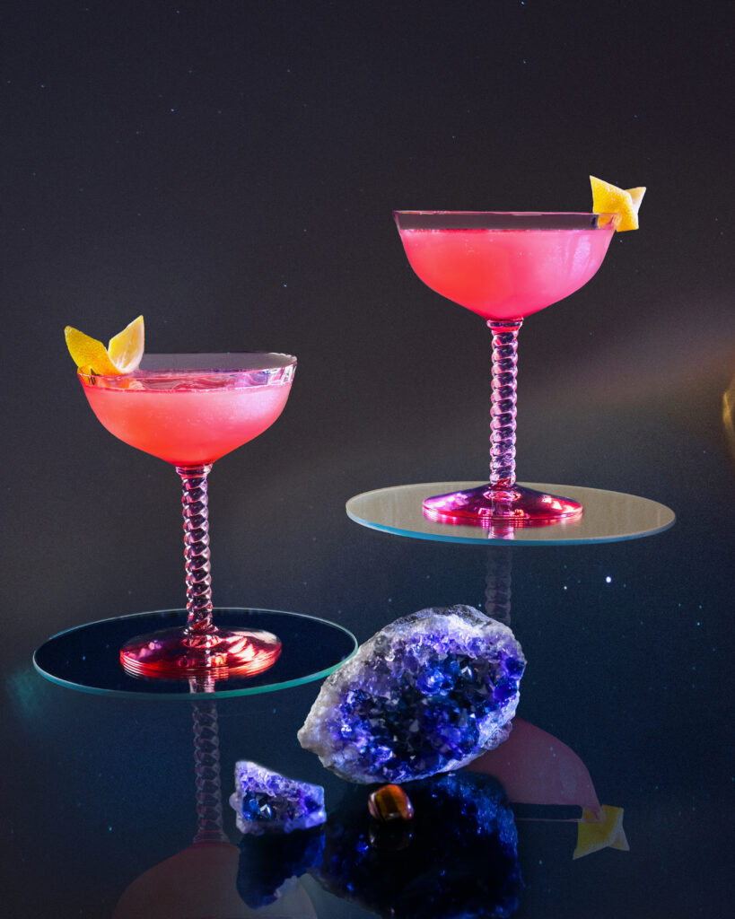 A cocktail for Libra includes tow glasses filled with a pink cosmopolitan with lemon garnish against a black background.
