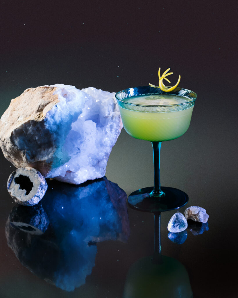A Corpse Reviver Cocktail for Pisces in a glass with green liquid and yellow garnishes all on a black reflective table.