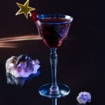 A Black Manhattan cocktail for Capricorn in a fancy glass with a star topper cocktail stick and crystals around the glass.