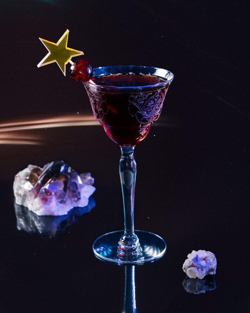 A Black Manhatten cocktail for Capricorn in a fancy glass with a star topper cocktail stick and crystals around the glass.