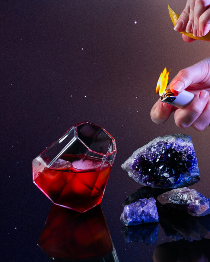 A geometric wine glass with red liquid inside as a cocktail for scorpio called scorched earth. A person holds a lit lighter nearby against the black background.