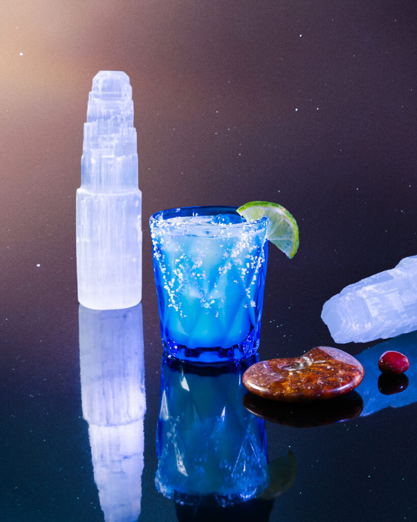 A margarita cocktail for cancer in a small blue glass with a lime garnish and salt rim. Selenite sits near by on the black table.