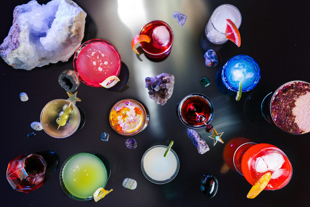 All 12 zodiac signs represented in a grouping of colorful cocktails in different shapes and sized of glasses with crystal on a black reflective surface, making the image look cosmic.