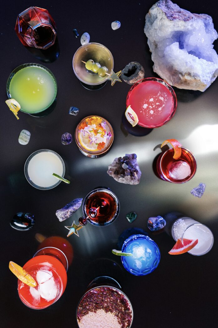 All 12 zodiac signs represented in a grouping of colorful cocktails in different shapes and sized of glasses with crystal on a black reflective surface, making the image look cosmic.