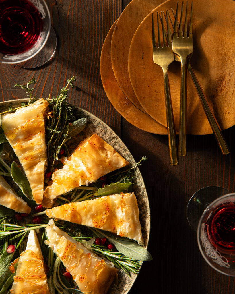 Triangle cuts of a phyllo pastry with fresh herbs as garnish in between, on a round plate in the left corner, and a stack of terra cotta colored plates with three gold forks on a wooden surface.