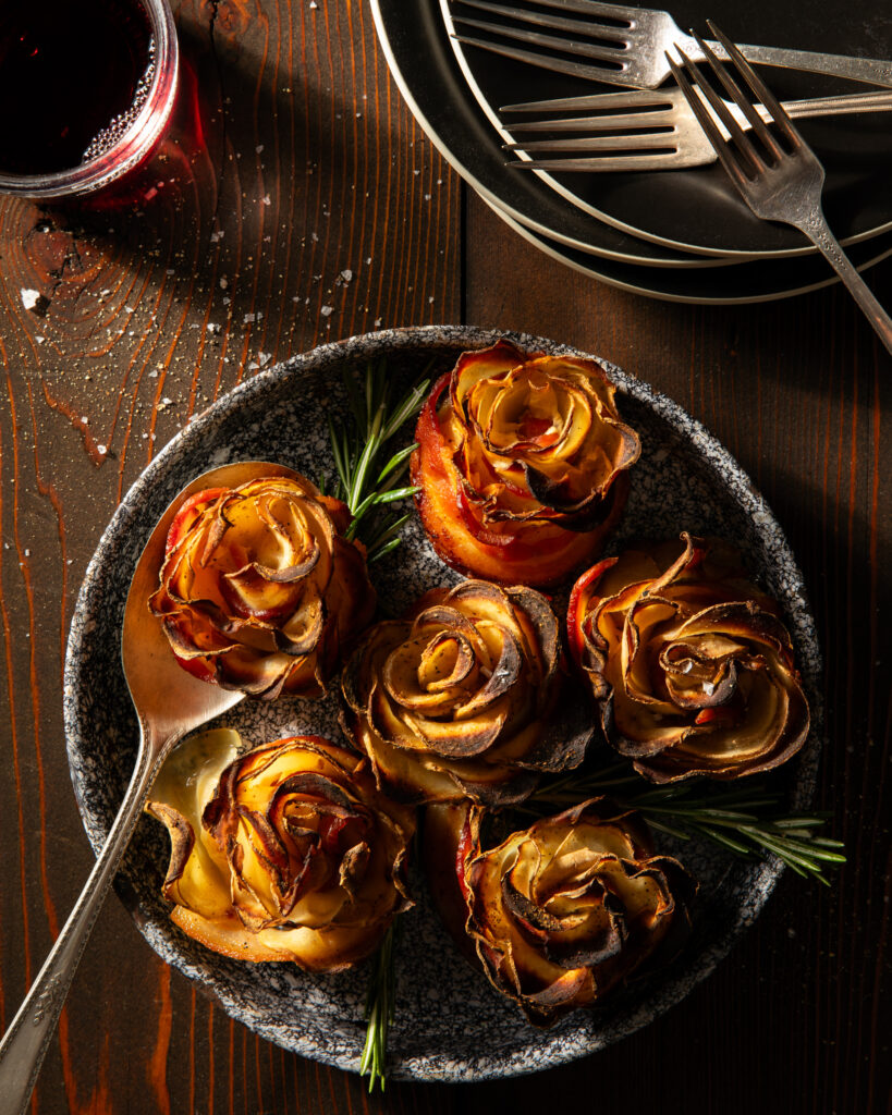 A round crackled looking plate with 6 bacon potato roses garnished with fresh rosemary, a glass or red wine and a stack of little plates and forks.