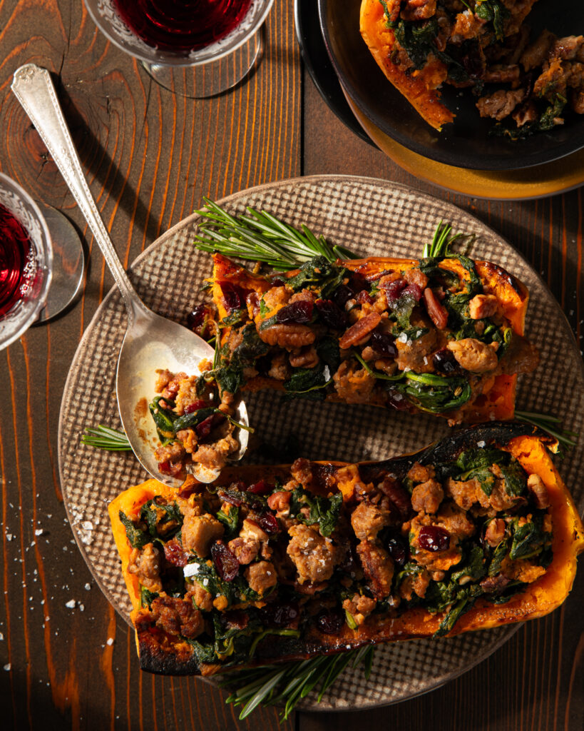 A textured plated on a wooden surface with 2 butternut squash halves, stuffed, a large silver serving spoon and rosemary garnish.