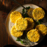 Seven round, yellow corn pudding cakes on a plate garnished with fresh herbs, sitting on a wooden surface.