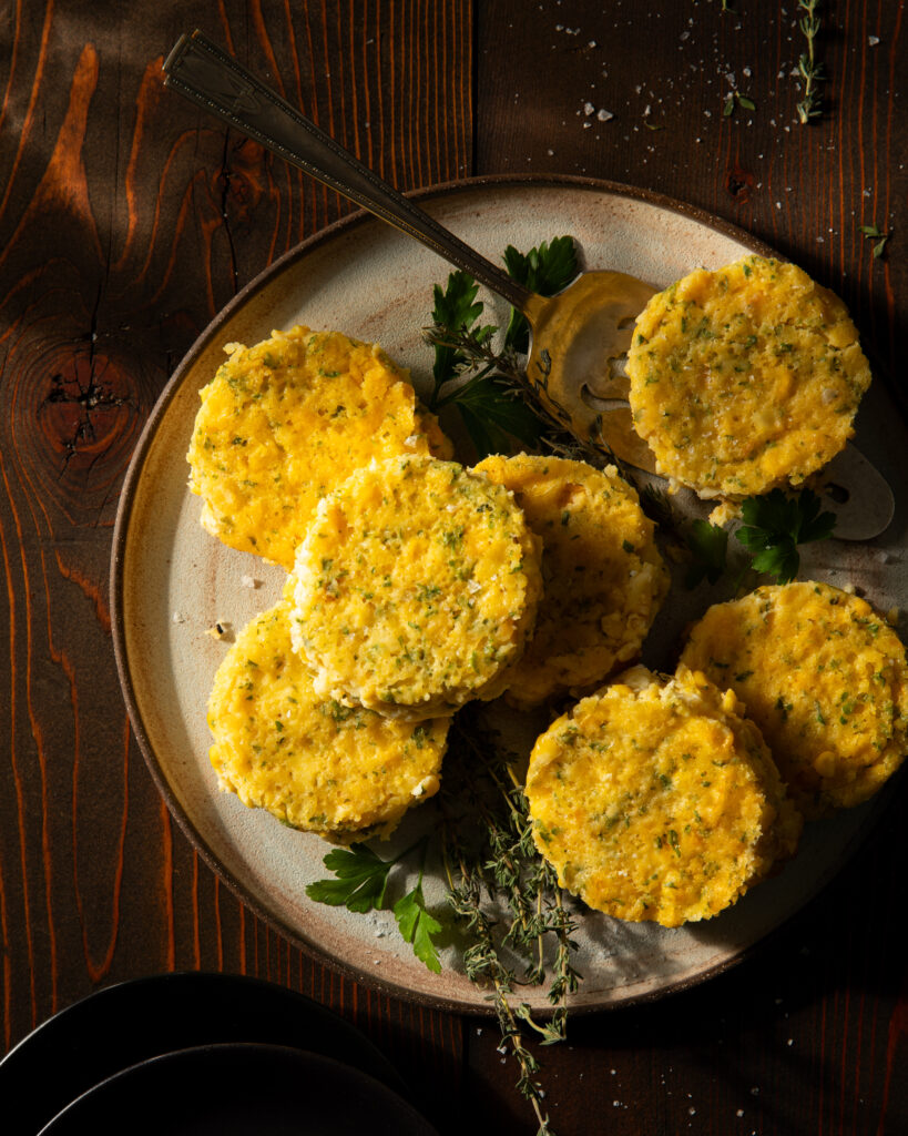 Seven round, yellow corn pudding cakes on a plate garnished with fresh herbs, sitting on a wooden surface.