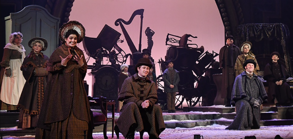 a group of stage actors play the villagers of A Christmas Carol onstage with instruments in the background.