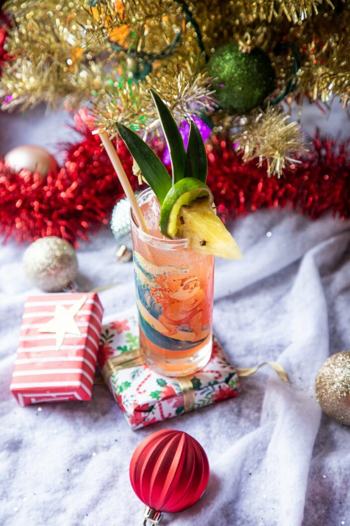 A tropical glass filled with a cocktail and topped with fresh fruit sits on top of a present surrounded by tinsel.