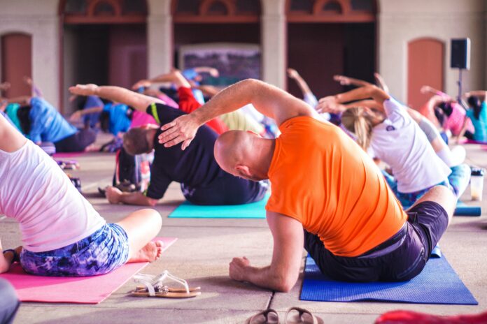 A group of people take part in a yoga class, stretching and sitting on colored yoga mats on the floor.