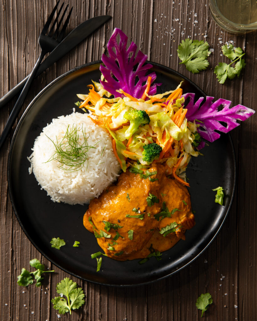 A black plate with coconut chicken, rice, and vegetables arranged on top from Kilimanjaro Flavour. Various herbs and colorful edible flowers are scattered throughout as well.