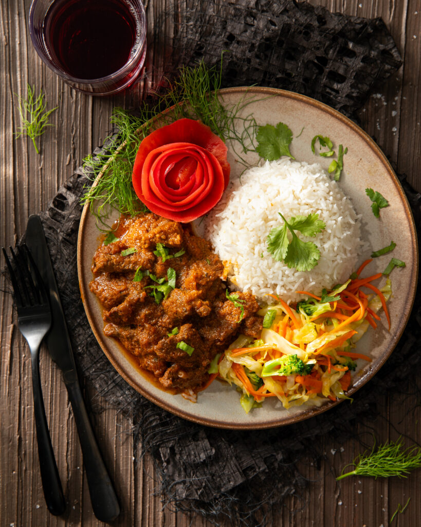 A cream colored plate with braised beef, rice, a mixed vegetable slaw, and an edible flower on top from kilimanjaro flavour.