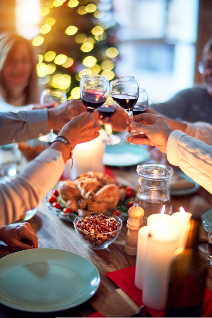Four people toast at a Holiday setting with holiday wine glasses over a table full of food and candles.
