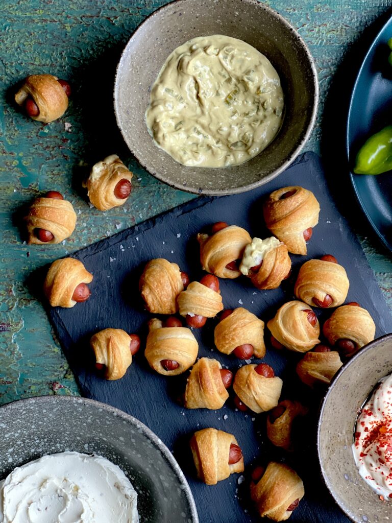 Mini pigs in a blanket on a slate tray sitting on a greenish surface with 3 bowls of dips.