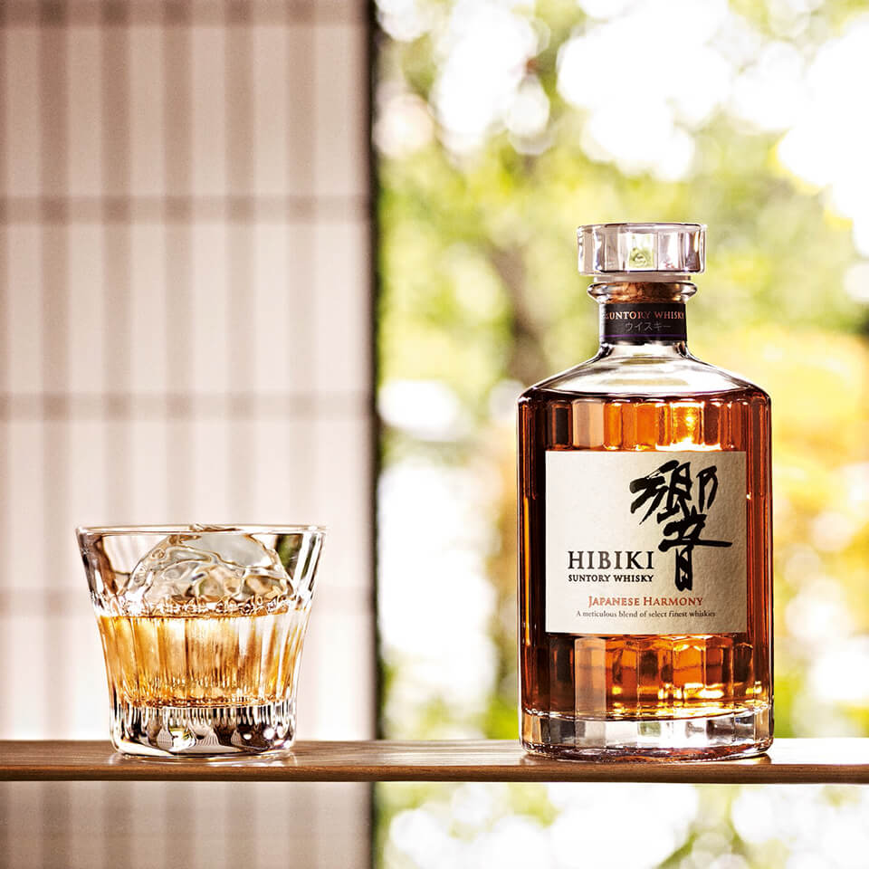 A bottle of Japenese whiskey sits on a table beside a whiskey glass filled with the drink and an ice cube, all in front of a tree background.