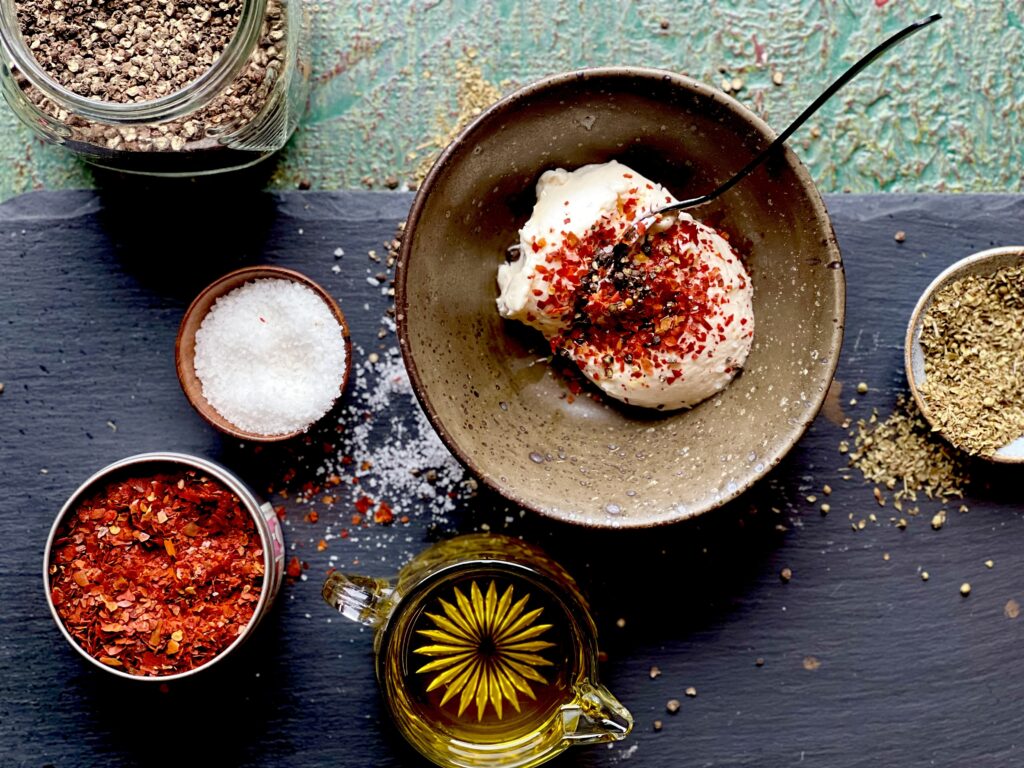 Bowls with seasonings and oil on a black slate surface surrounding a brown bowl of harissa chili mayo dip.