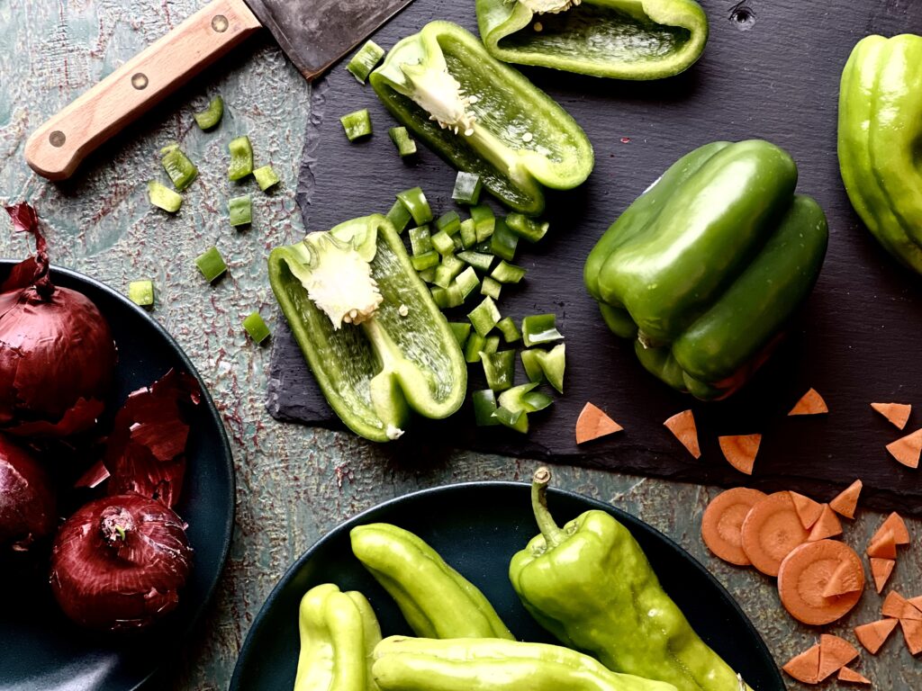 Fresh green peppers, some whole and some chopped, carrots chopped in little triangles, and whole red red onions on cutting boards and small plates on a grey background with part of a large chef knife showing in the upper left corner.