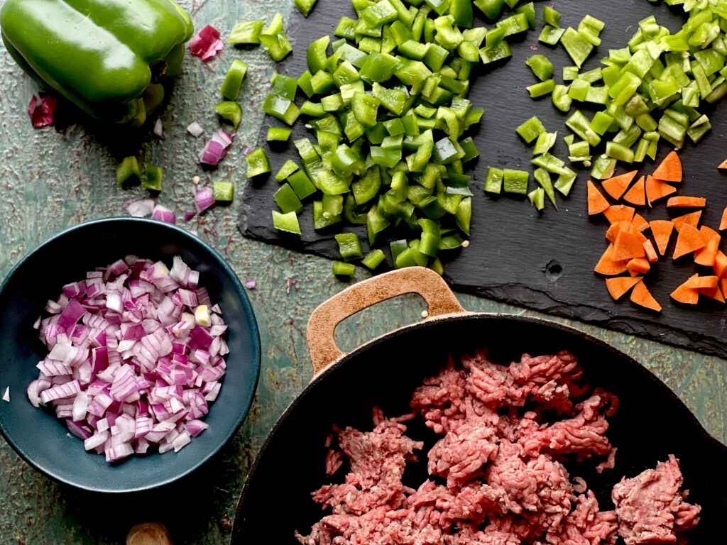 Minced red onion, green pepper and carrots on a slate surface and in a bowl with a cast iron skillet with raw ground beef.