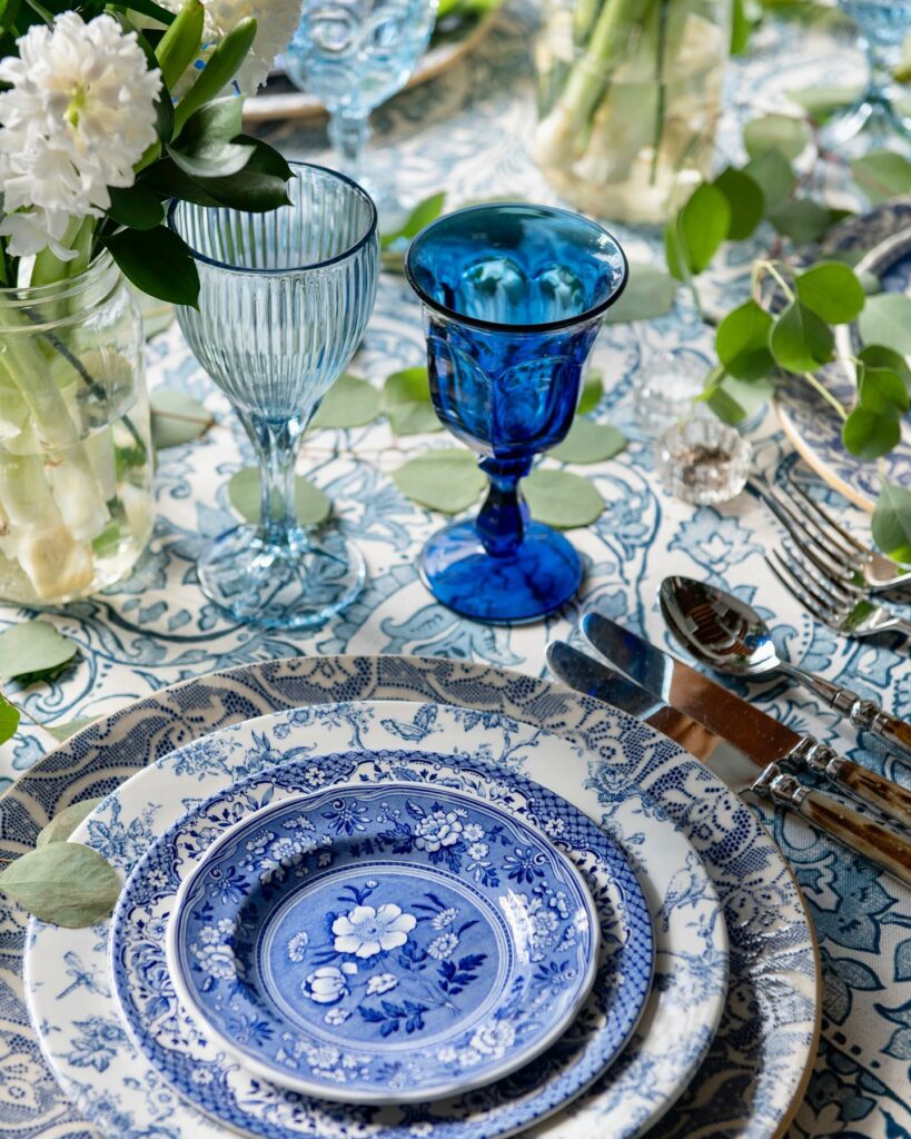 A stack of blue floral patterned plates sit beside opaque blue glasses, silverware, and other decorations.
