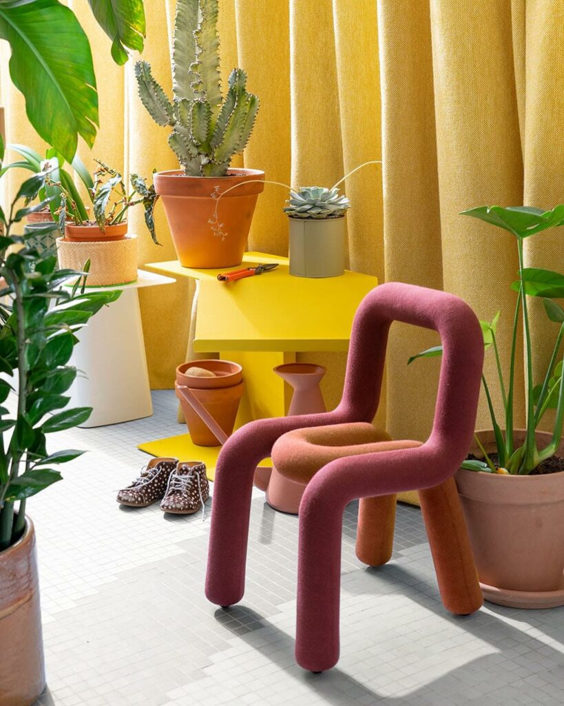 A red chair made of tubing sits in the center of many different green plants in front of a yellow curtain and table at Moustache design shop in Paris