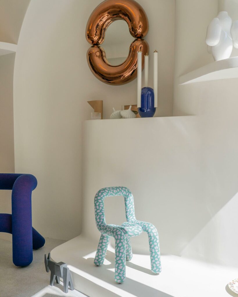 A light blue tube chair sits in the middle of a white room with a balloon tube mirror on the wall and dark blue tube table to the left at Moustache in Paris.