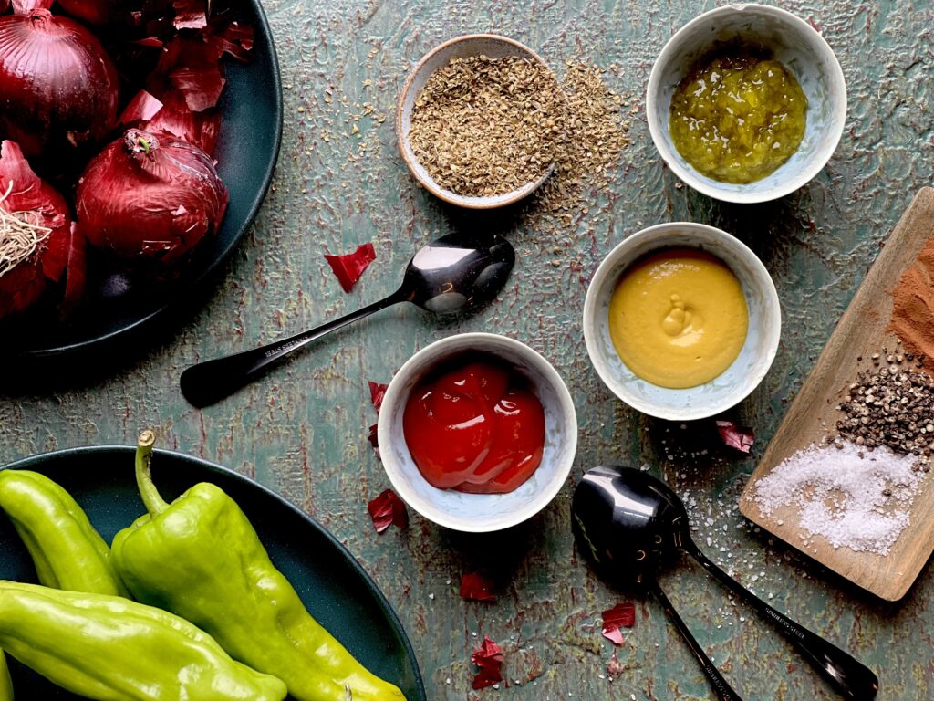 Four small bowls, 1 with seasoning, 1 with relish, 1 with mustard, and one with ketchup on a blue/grey surface with a small wooden tray on the right with salt, pepper and paprika, and fresh whole red onions and habanero pepper s on the left. 