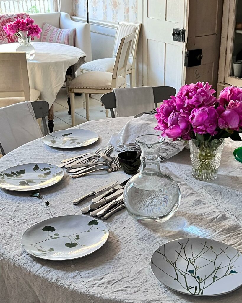 A vintage white table cloth covers a table with simple floral white plates, silverware, a jug of water, and purple flowers in the center of the table.