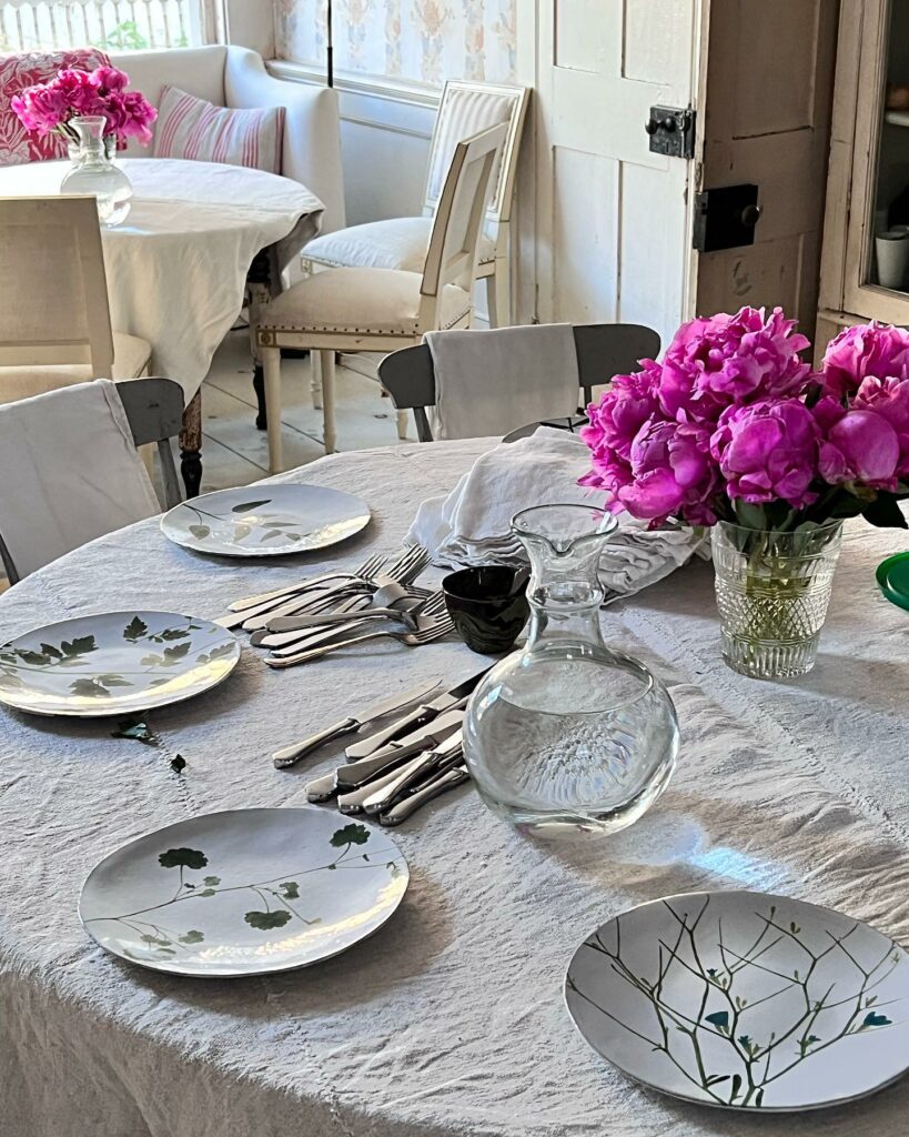 A vintage white table cloth covers a table with simple floral white plates, silverware, a jug of water, and purple flowers in the center of the table.