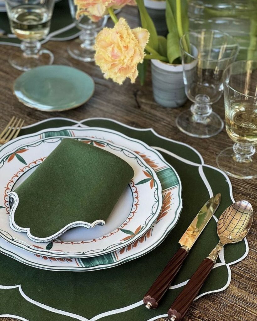 A wooden table holds a green themed plate setting with a green and white napkin, green accented plate, dark green placemat, and glasses surrounded the setting.