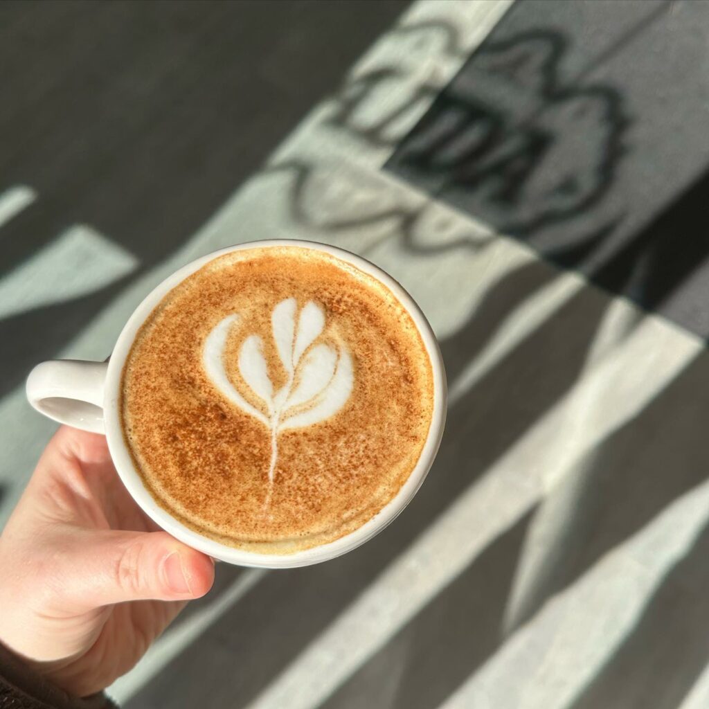 A person holds a coffee mug filled with a latte and topped with a leaf design in front of the shadow of Adda Coffee and Tea in Pittsburgh