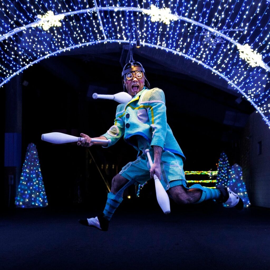 A man in a silly costume jumps in the air in front of lights while juggling bowling pins.