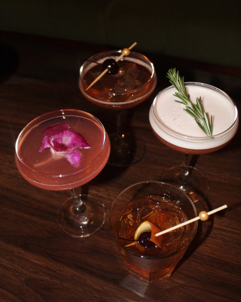 Four cocktail glasses filled with different pink and white cocktails topped with stick garnishes at PA Market in Pittsburgh.
